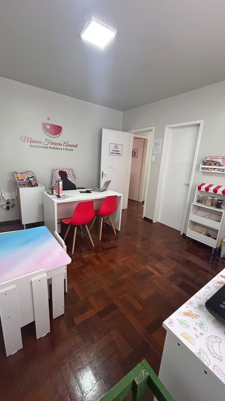 Consultório com mesa branca, cadeiras vermelhas e prateleiras com brinquedos